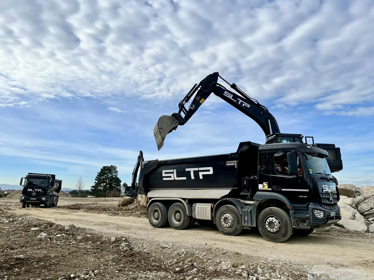 Pelle noire chargeant un camion-benne noir, tous deux siglés SLTP, sur le chantier Vectura, à Rillieux-la-Pape.